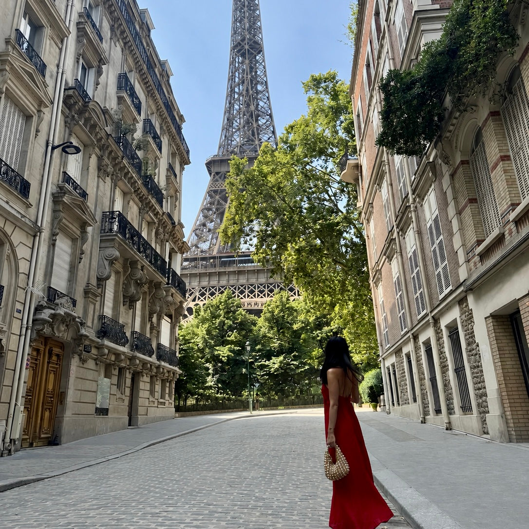 A woman in a long red dress stands on a cobblestone street in Paris, holding a woven cream ZOEL bag with pearls and looking toward the Eiffel Tower, framed by classic beige buildings and green trees under a blue sky.