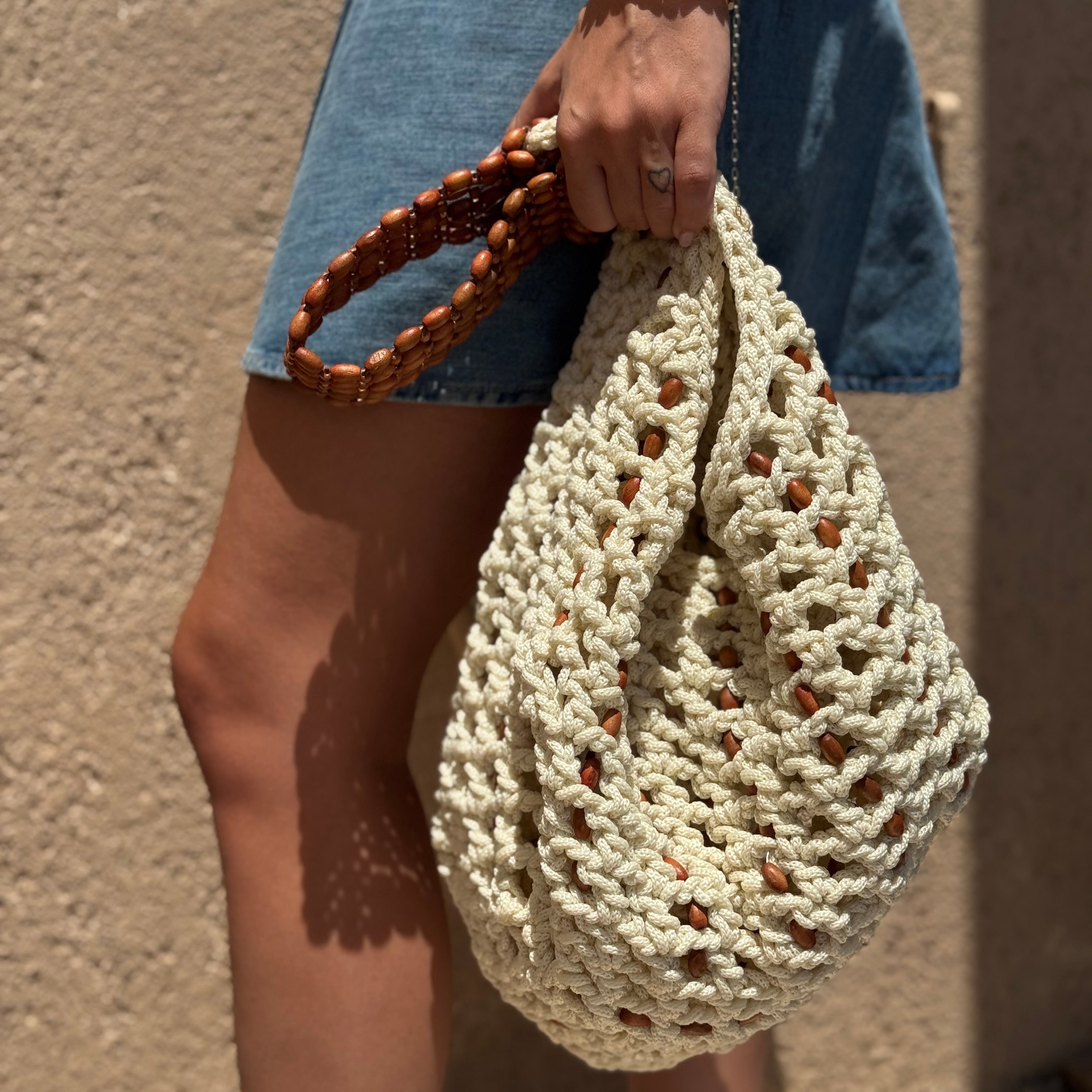 A person wearing a denim skirt holds a chunky cream crocheted shoulder bag with a brown woven handle, standing against a textured beige wall.