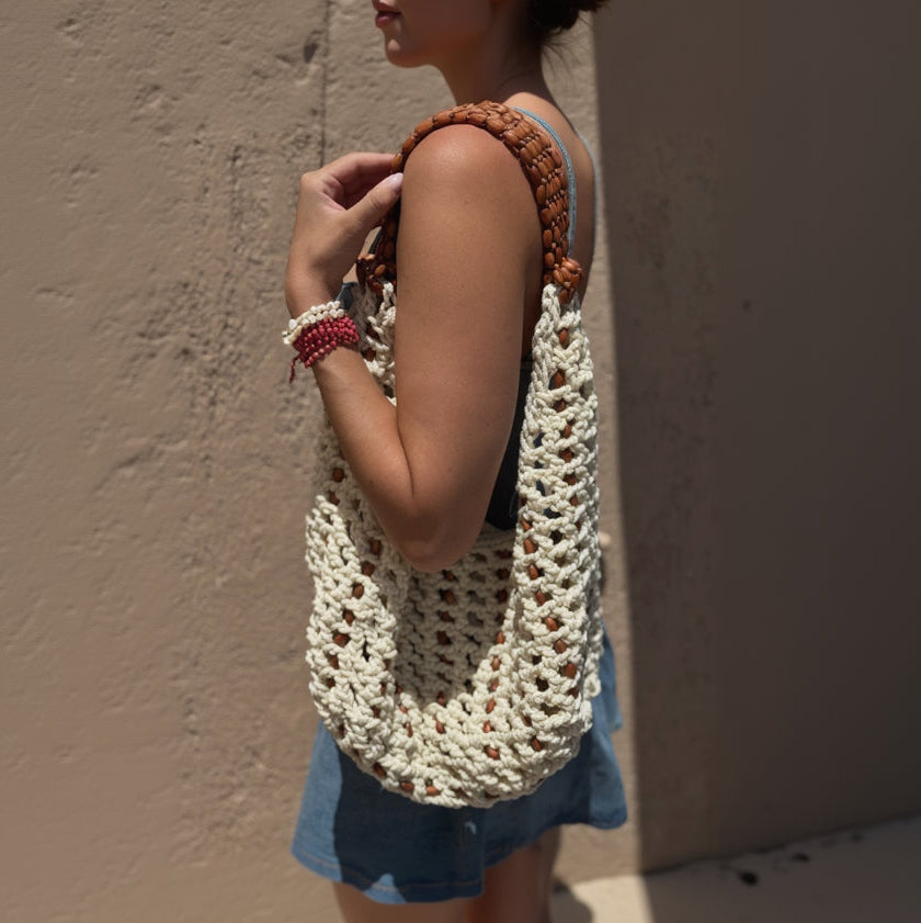 A close-up lifestyle shot of a person wearing a chunky, open-knit crochet shoulder bag in cream with brown wooden bead straps, set against a textured beige wall.