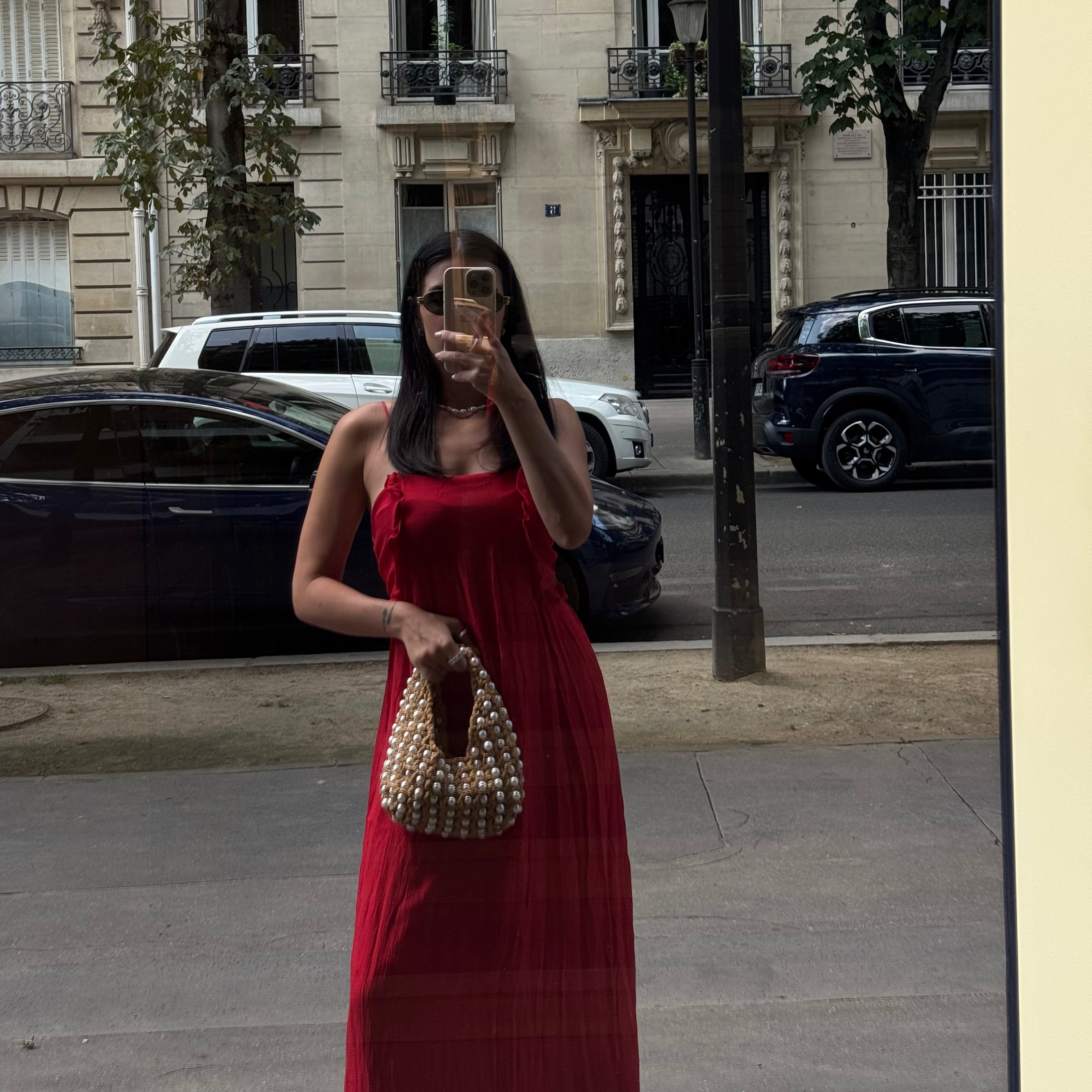 A woman takes a mirror selfie in an urban street setting, wearing a sleeveless red maxi dress and holding a pearl-beaded handbag made by ZOEL.