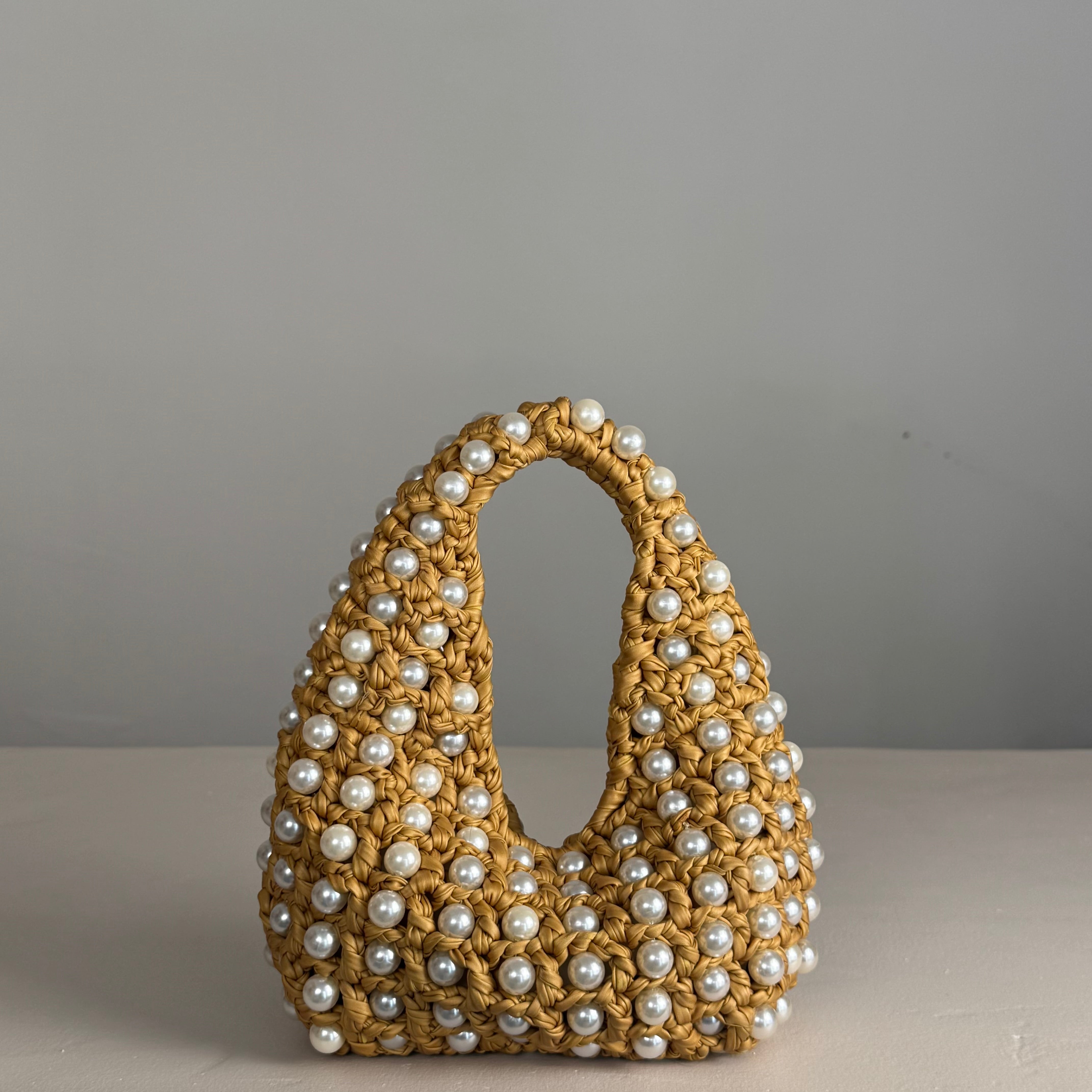 A woven straw handbag with a loop handle, heavily embellished with white faux pearls, set against a neutral gray background.