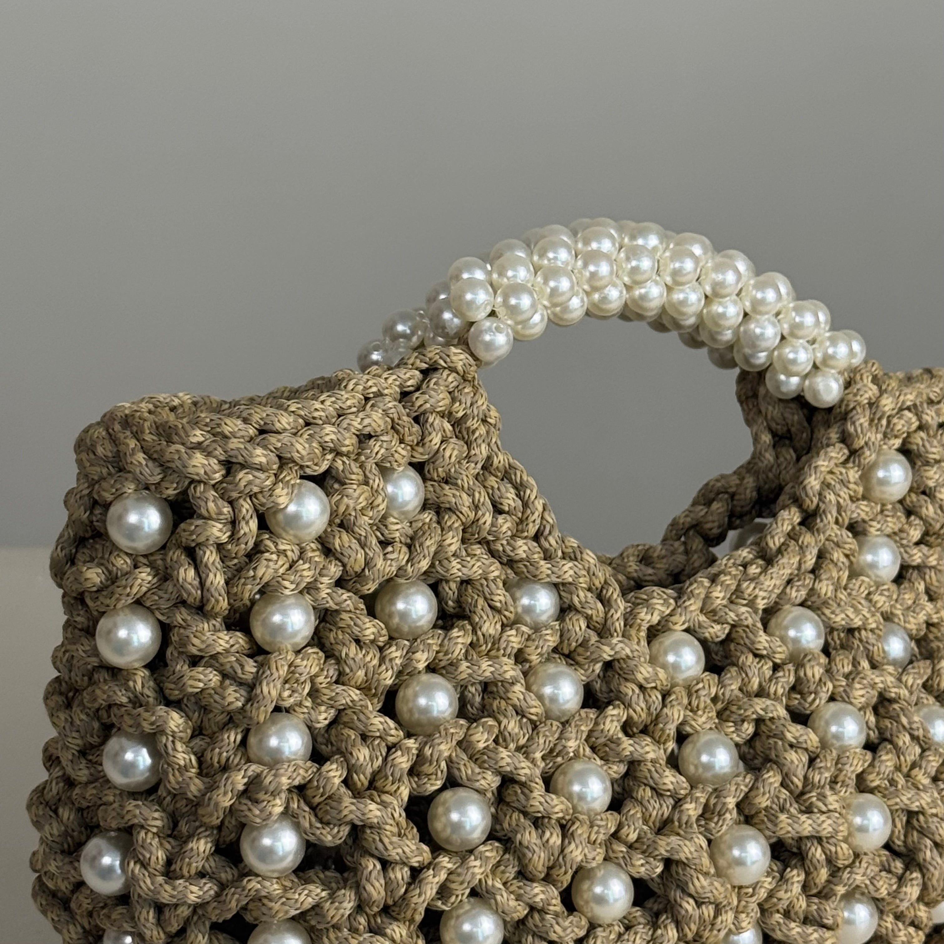 A close-up of a chunky crocheted handbag in taupe with white faux pearls woven throughout and a pearl-trimmed handle, set against a neutral gray background.