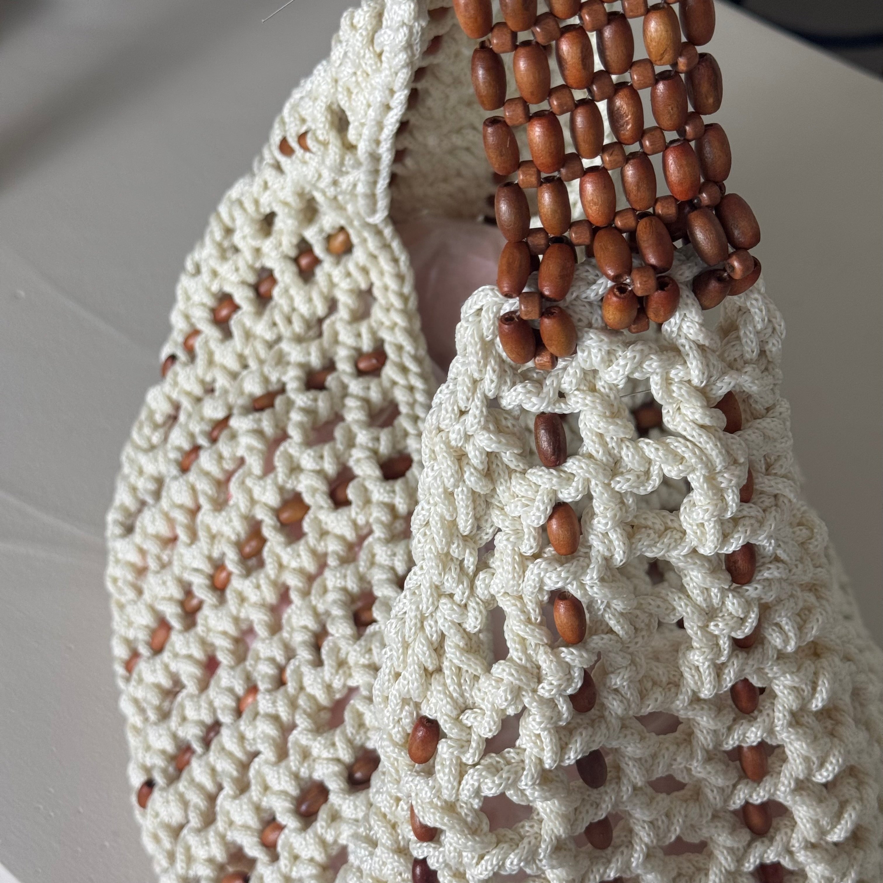 Close-up of a handmade crochet shoulder bag with a woven wooden bead handle, set against a neutral light gray background.
