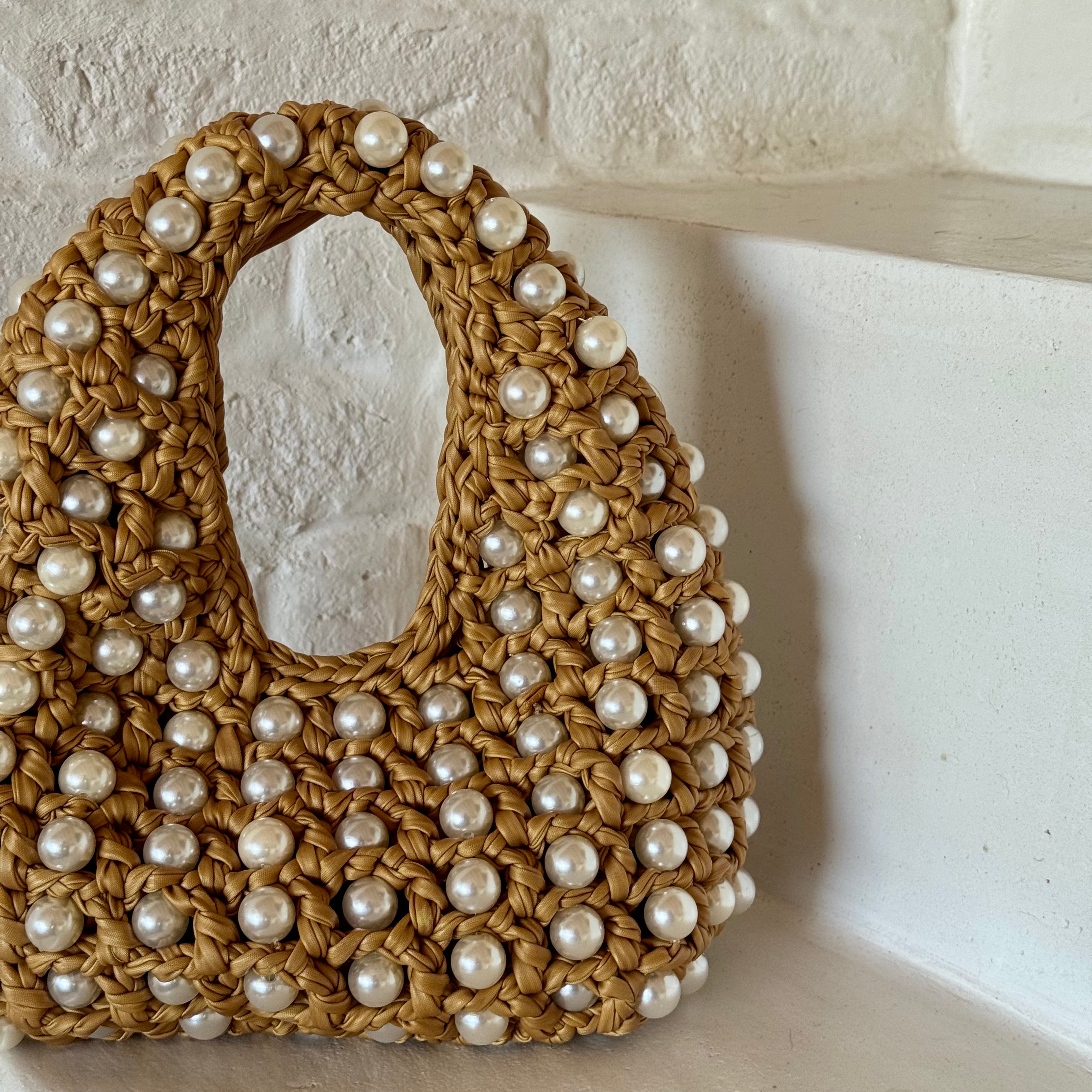 A woven straw shoulder bag with a large oval cutout handle, decorated with numerous white faux pearls, set against a white brick wall background.