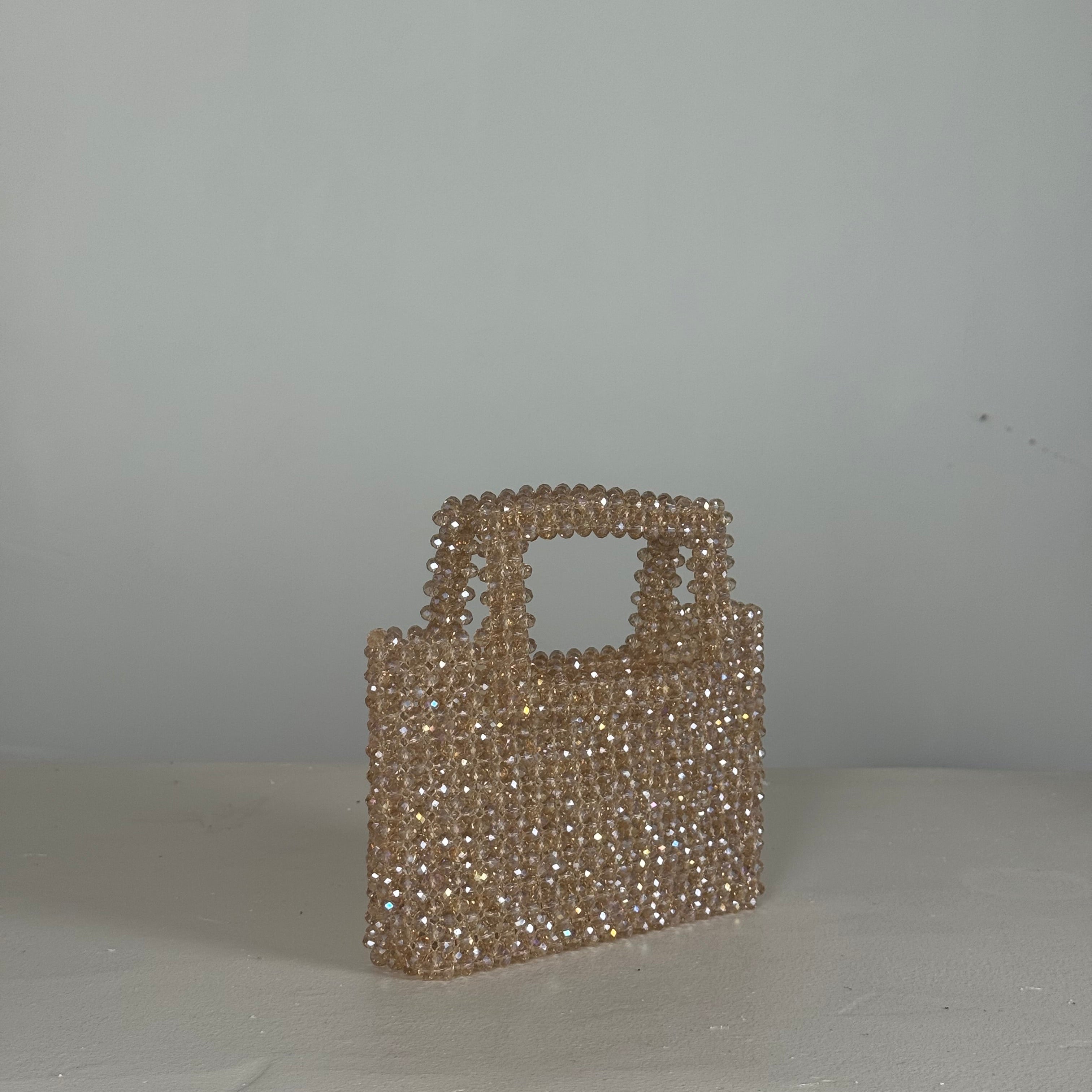 A small structured handbag covered in clear rhinestones, photographed hanging against a plain light gray wall.