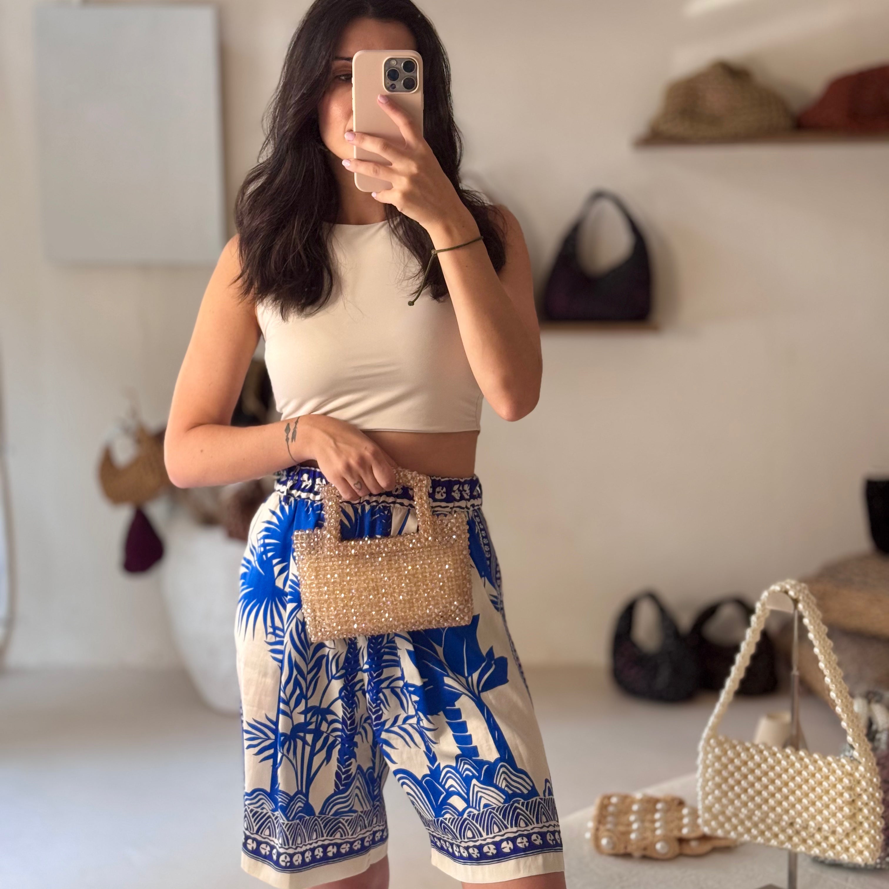 A woman takes a mirror selfie in a boutique, wearing a beige sleeveless crop top and high-waisted shorts with a blue and white palm tree pattern. She is holding a small woven beige handbag.