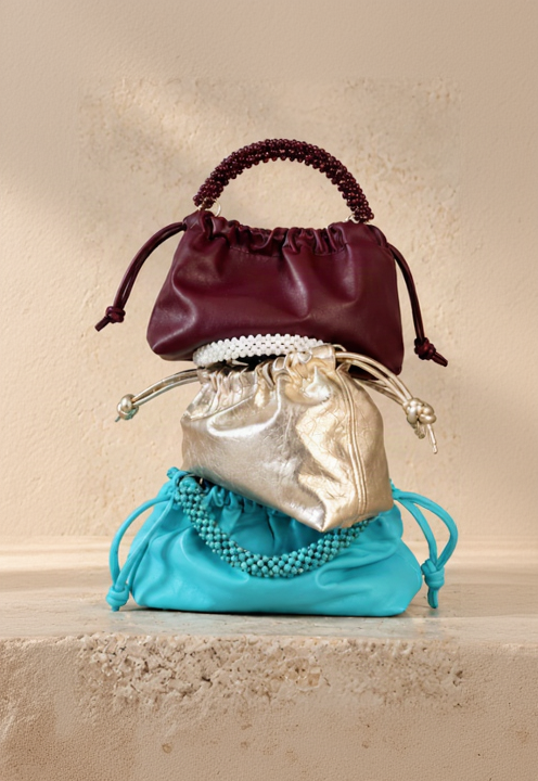 Three small drawstring handbags stacked on a stone ledge against a textured beige wall with leaf shadows. The bags are burgundy, metallic gold, and turquoise blue.