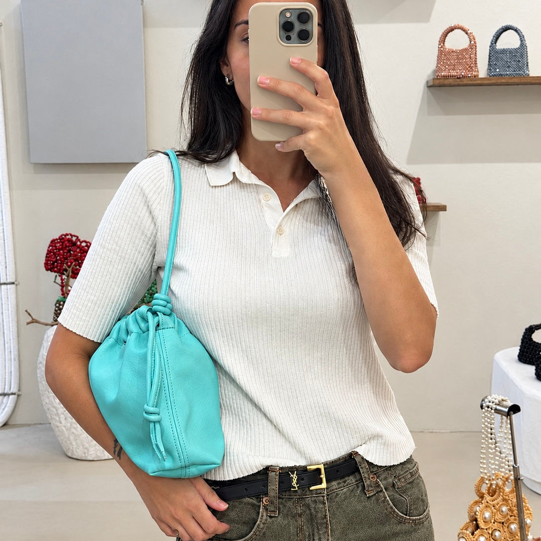 A woman takes a mirror selfie in ZOEL showroom while wearing a white ribbed short-sleeve polo shirt and olive green jeans, carrying a bright turquoise drawstring pouch bag from ZOEL.