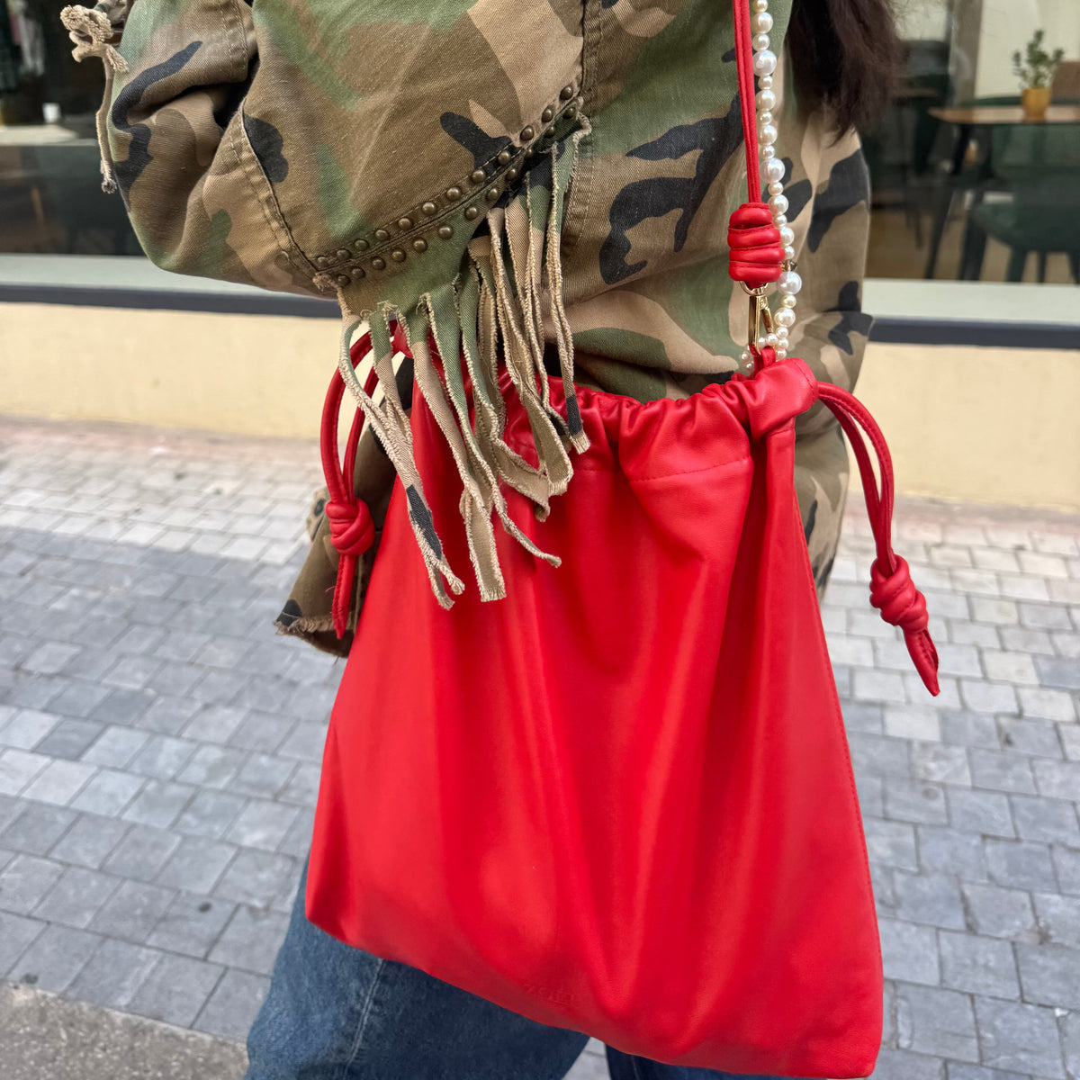 A person outdoors wearing a bright red drawstring shoulder pouch bag with a red cord strap and white faux-pearl bead detail, over a camouflage jacket.