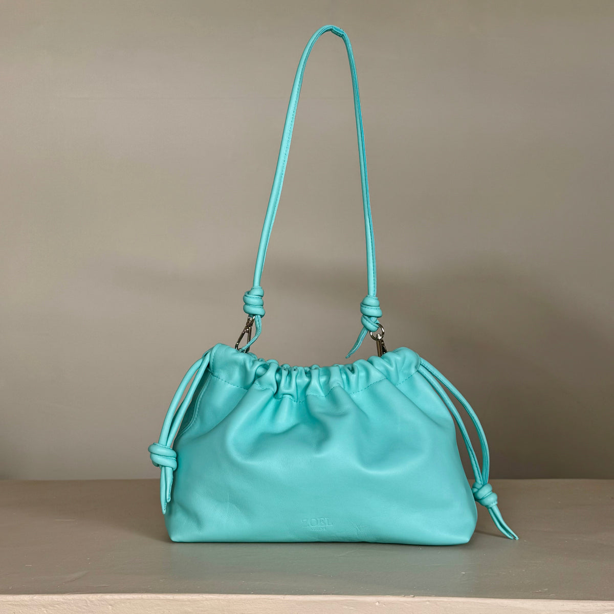 A turquoise leather shoulder pouch bag from ZOEL with a drawstring top and knotted strap details, displayed on a light beige surface against a neutral gray background.
