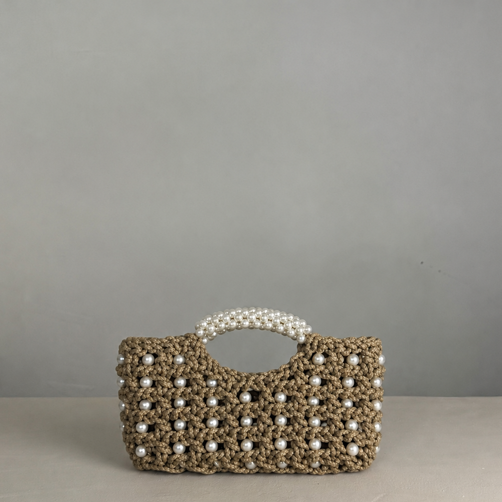 A crocheted beige handbag with white pearl accents and a pearl handle, displayed on a light gray surface against a gray background. Kimolos pearl crochet bag by ZOEL