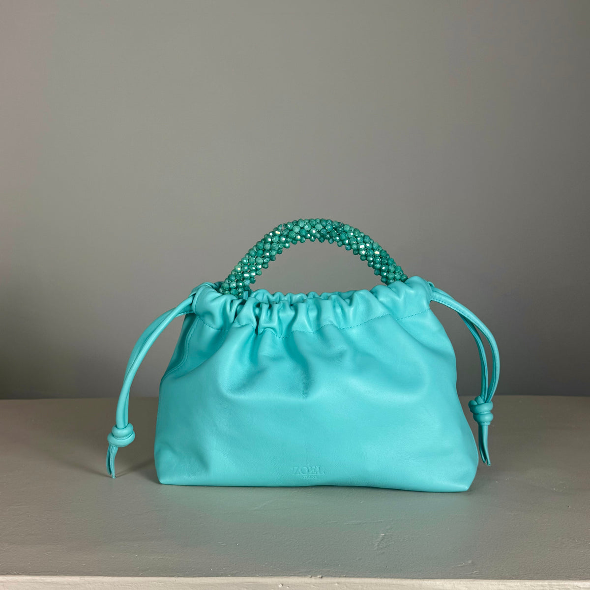 A turquoise leather drawstring pouch bag from ZOEL with a beaded top handle, displayed on a neutral gray surface against a gray background.