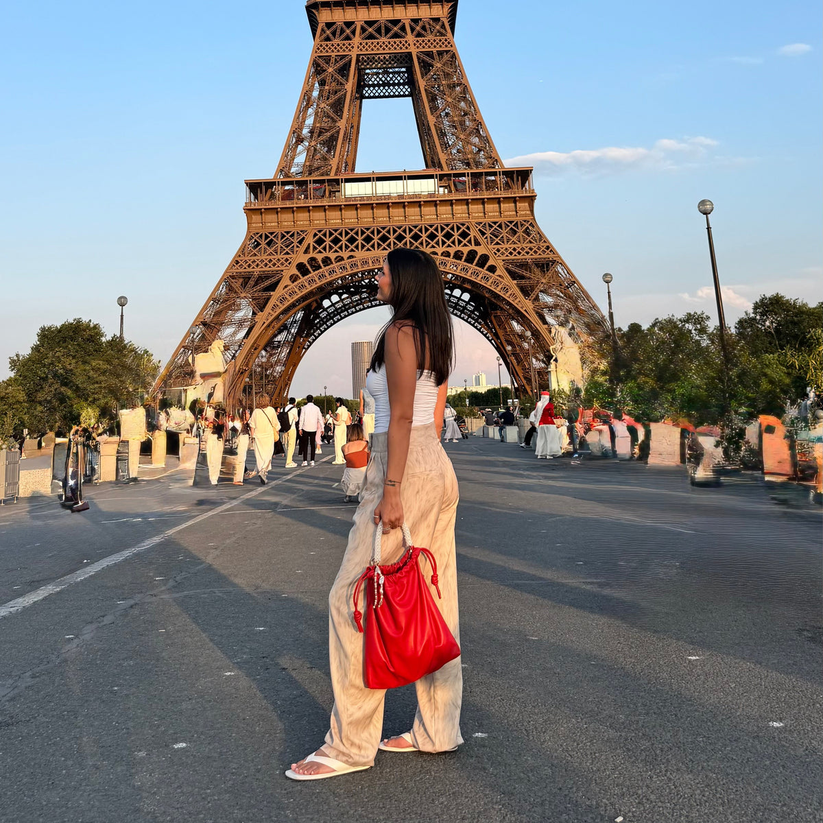 A woman in a white sleeveless top and beige pants stands in front of the Eiffel Tower, holding a bright red pouch bag with white crystal handle from ZOEL.