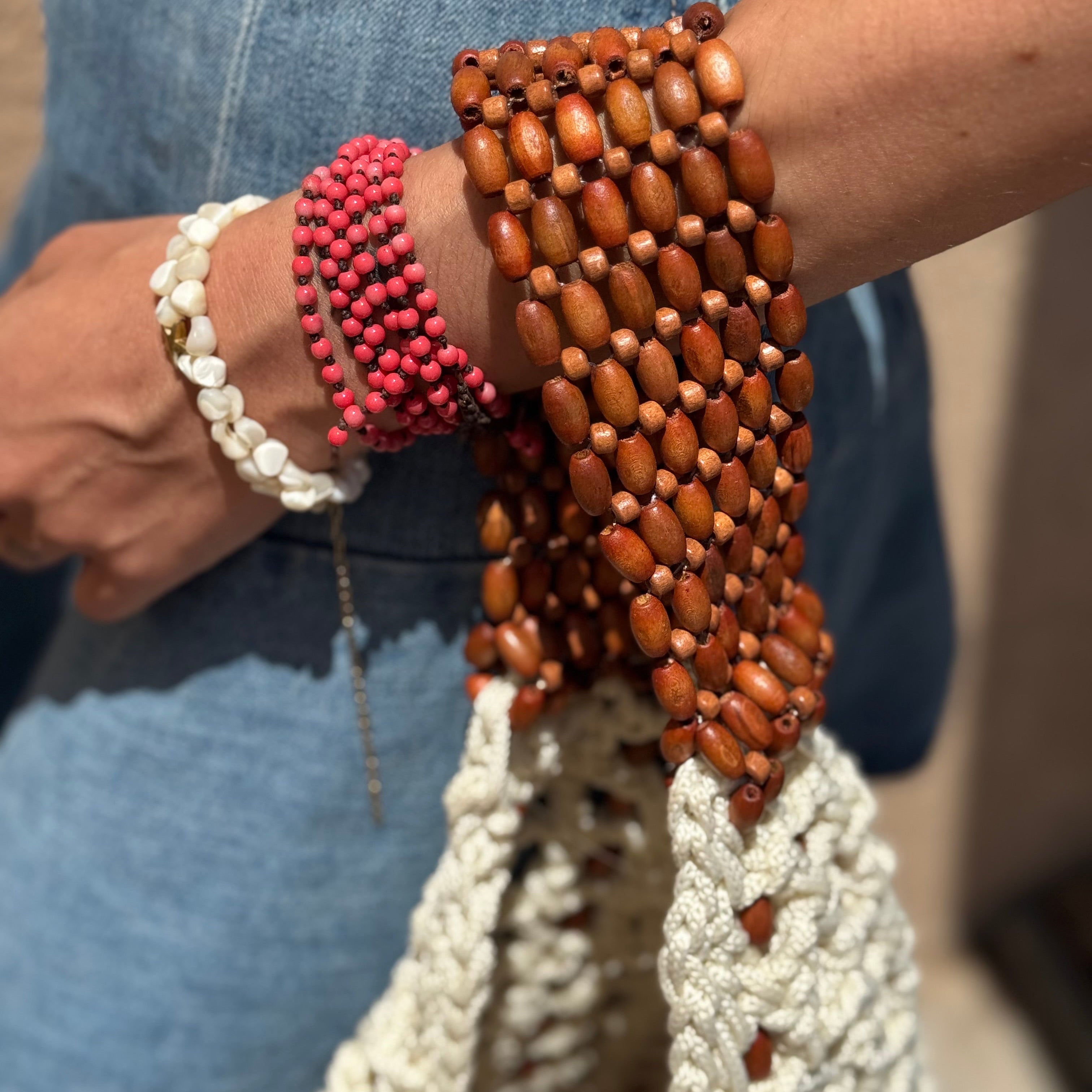 A close-up of a person's wrist and hand wearing multiple beaded bracelets, including a wide brown wooden bead wrap, a pink beaded bracelet, and a white beaded bracelet, paired with a chunky white crochet bag and blue denim clothing.