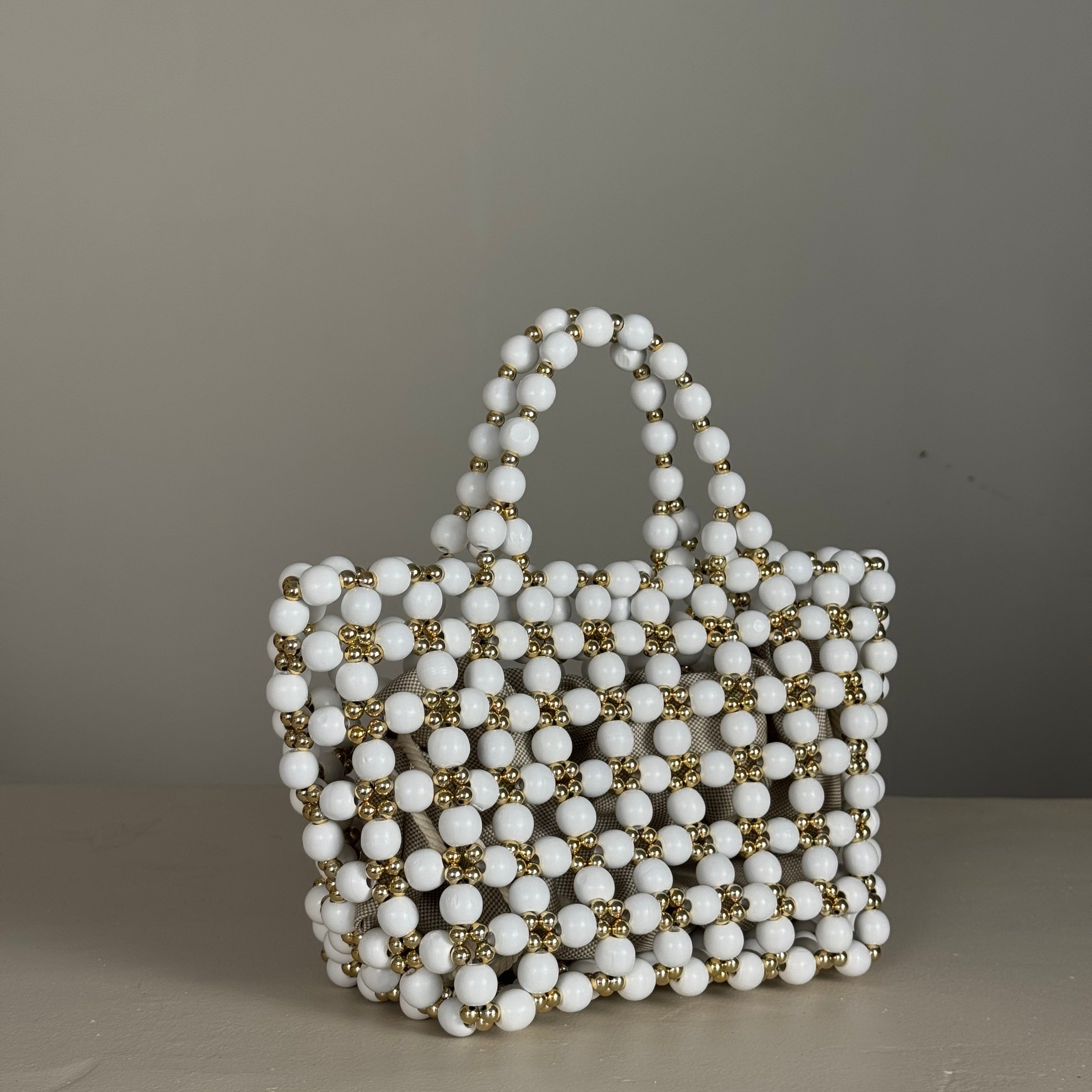 A structured handbag made of wooden white bead with small gold metallic accents, photographed hanging against a plain gray wallMade by Zoel