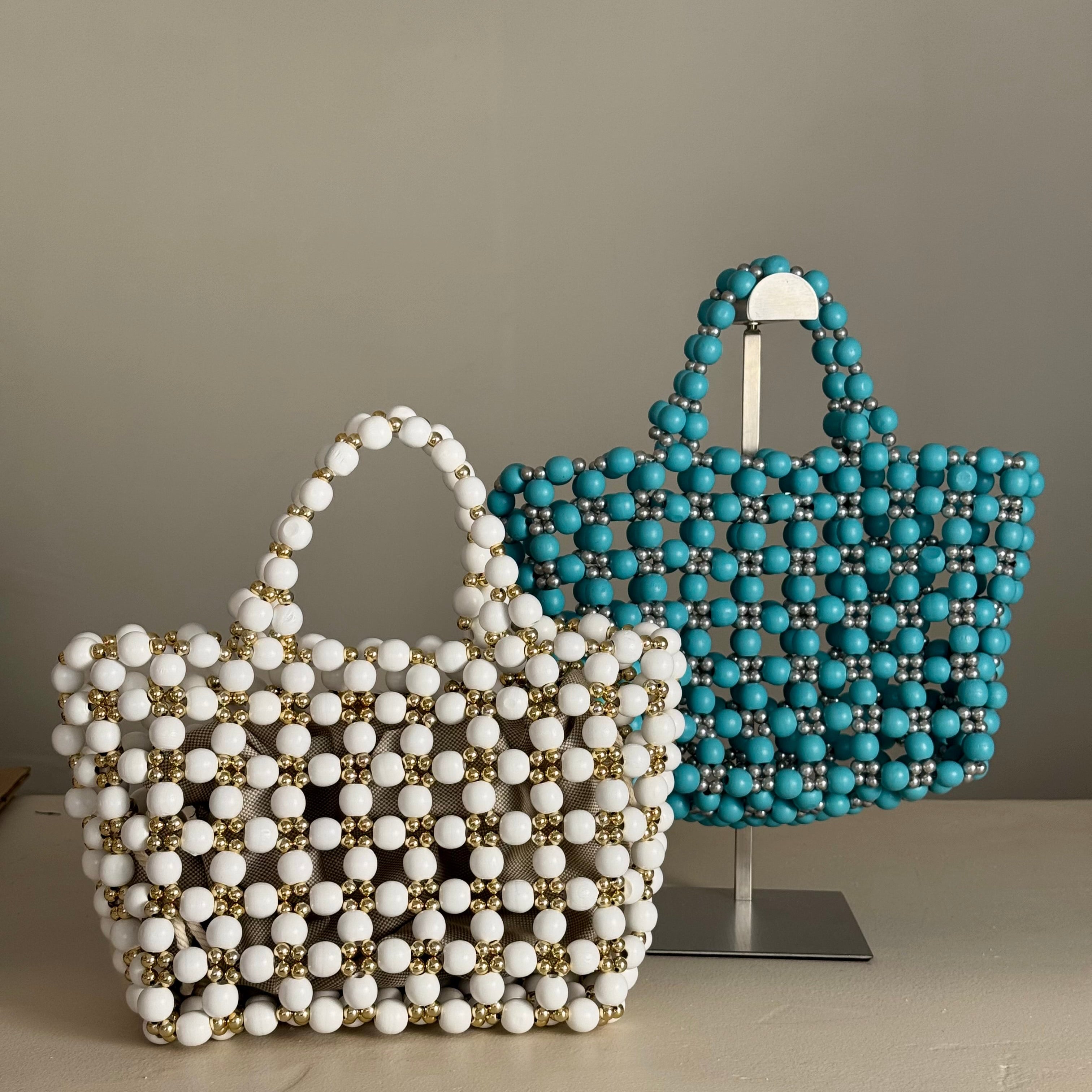 Two beaded wooden tote bags displayed on a stand against a neutral gray background. The bag in the foreground is white with gold accents, and the bag behind it is turquoise blue with silver accents. Made by Zoel