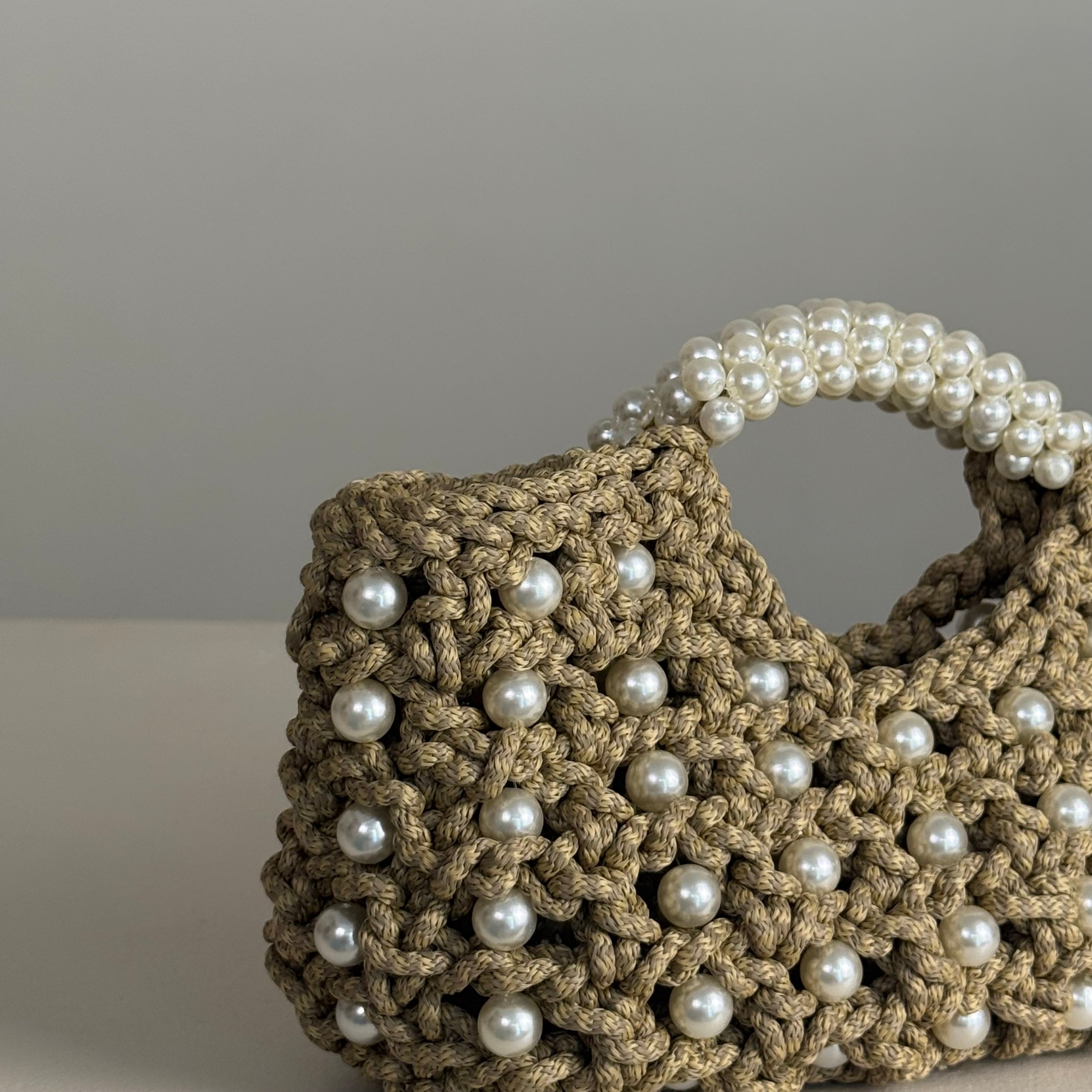 A close-up of a crocheted beige handbag with white faux pearls on the handle and scattered across the front, set against a neutral gray background.