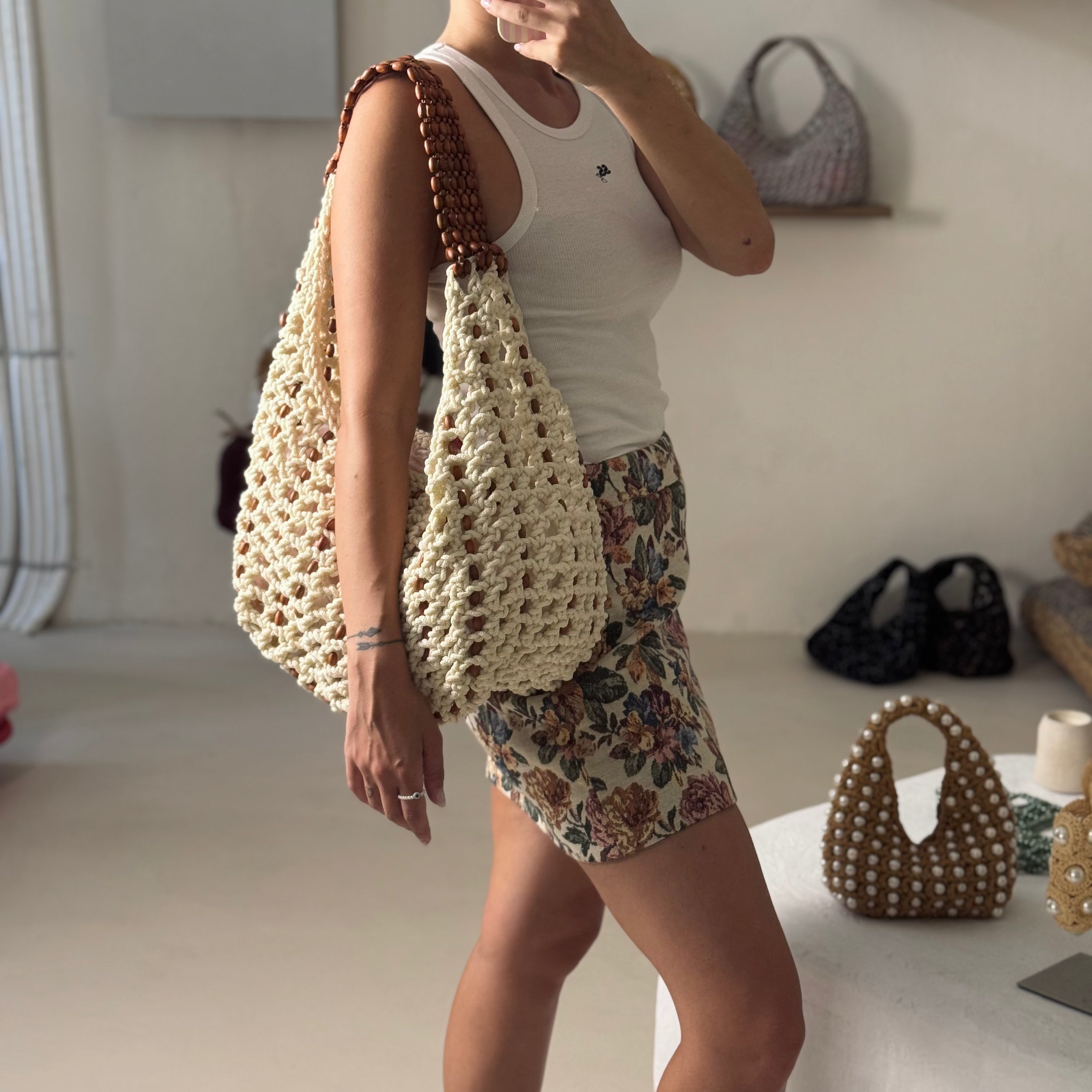 A woman wearing a white tank top and floral shorts carries a large cream crocheted shoulder bag with brown wooden beads on the strap in a bright indoor room.