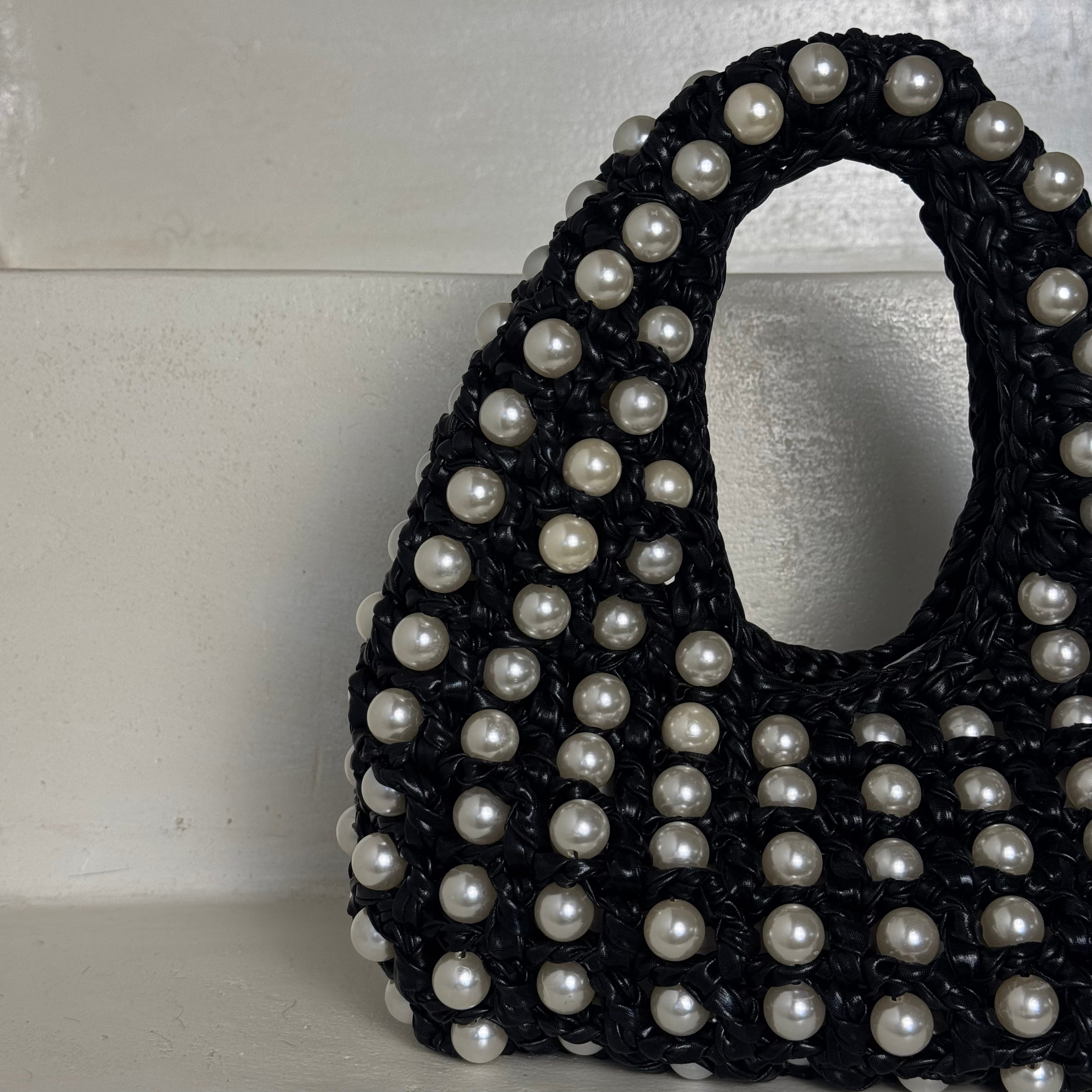 A close-up of a black woven or crocheted handbag covered with white faux pearls, photographed against a light off-white textured background. Made by Zoel