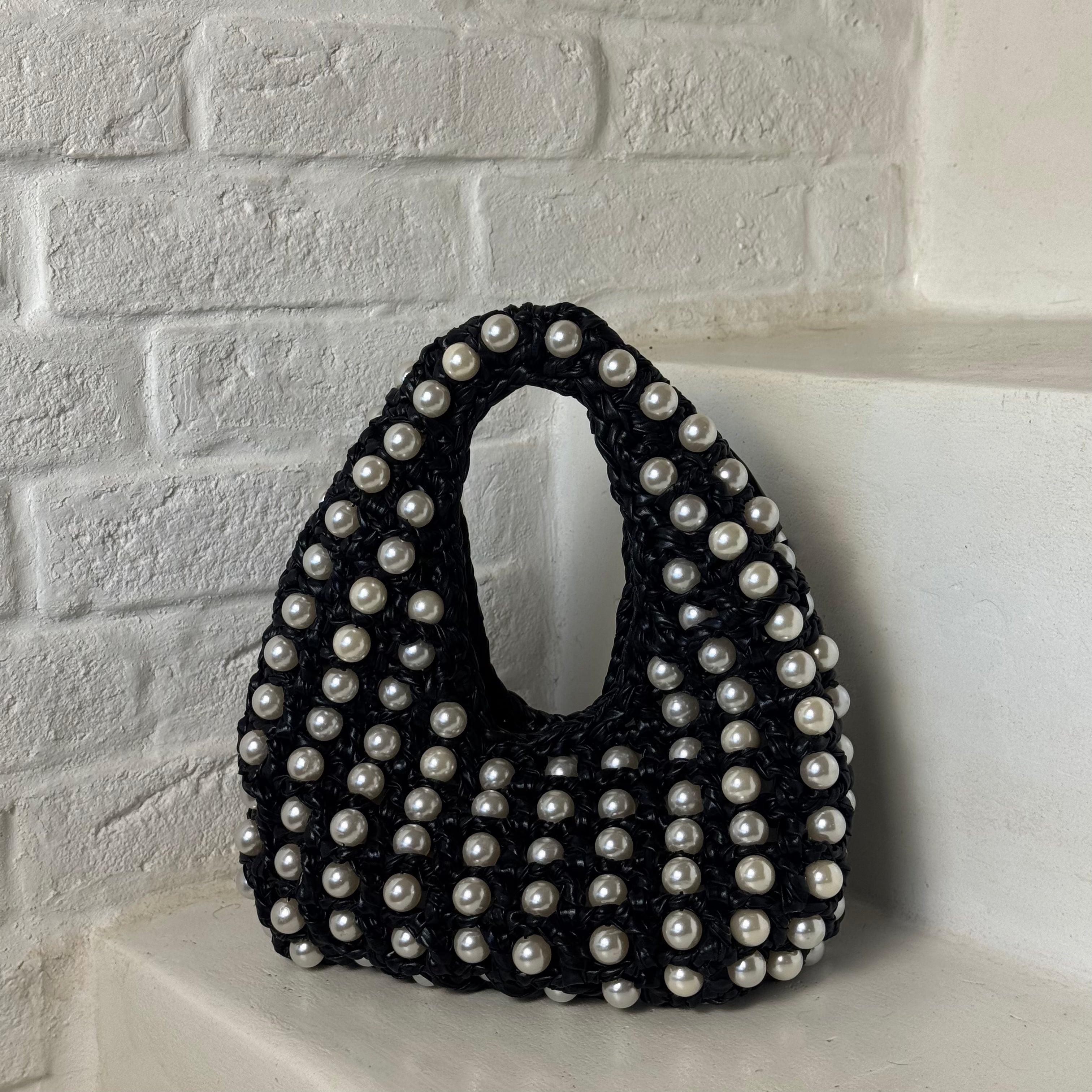 A black crocheted handbag adorned with white faux pearls, displayed on a white brick shelf.