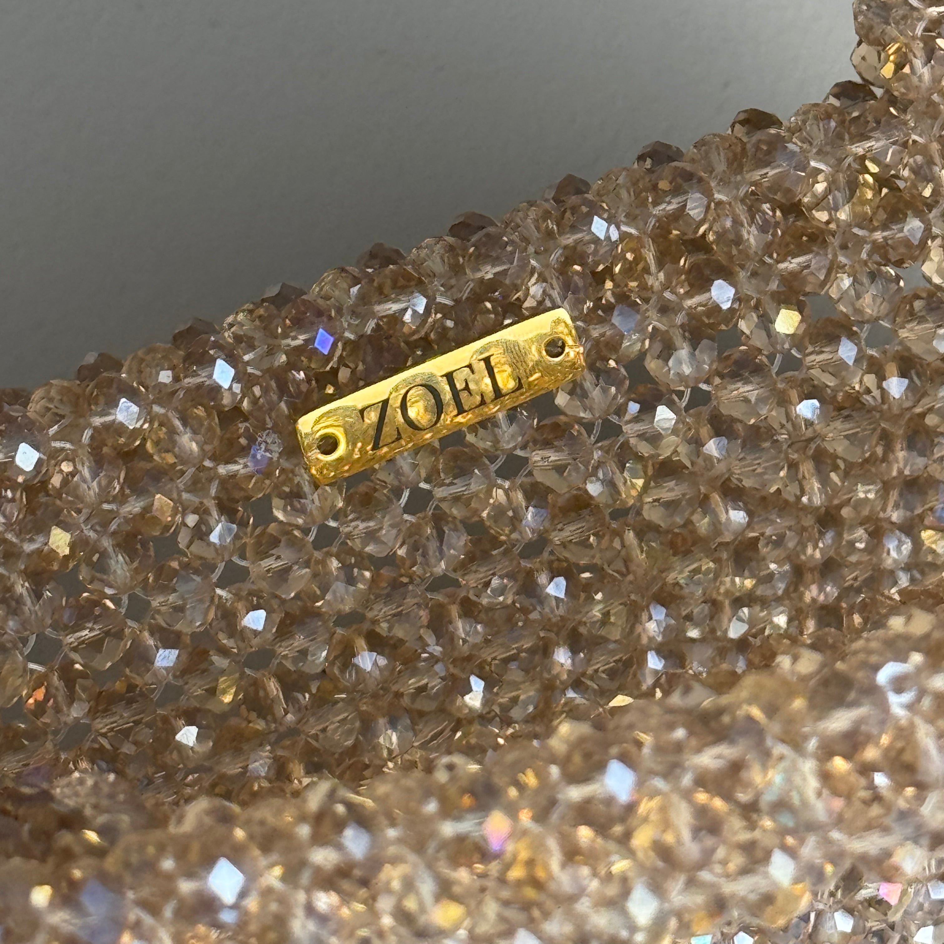 Close-up of a gold-tone metal tag engraved with 'HEIDI' attached to a heavily embellished, clear faceted crystal body with iridescent shimmer.