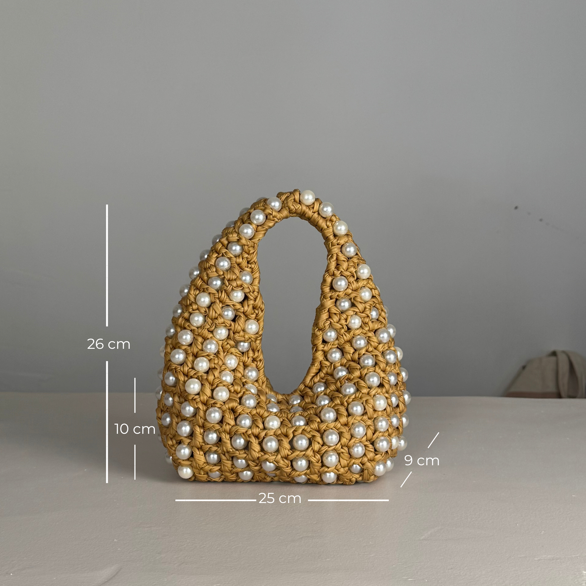 A crocheted mustard-yellow handbag covered with white faux pearls, shown on a light gray surface with overlaid dimension lines and measurements (26 cm height, 10 cm depth, 9 cm width, 25 cm base). Made by Zoel